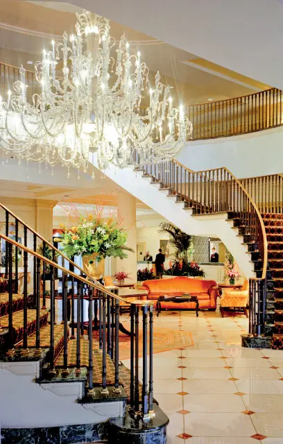 Grand foyer with a large crystal chandelier, a curved staircase with patterned carpet, marble floors, and an orange sofa