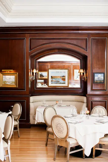 Dining room with dark wood paneling, upholstered banquette seating, and tables set with white linens and chairs.