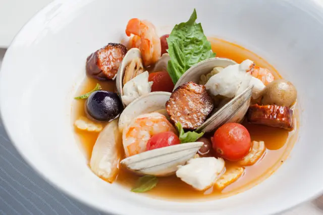 Close-up of Frogmore Stew with shrimp, clams, crab meat, sausage, tomatoes, olives, and basil in a white bowl.