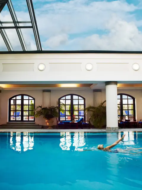 Indoor swimming pool with bright blue water, a person swimming, arched windows with dark frames, and a glass ceiling