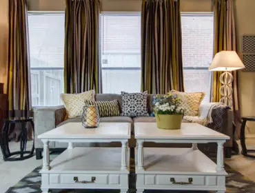 Living room with a gray sofa, patterned throw pillows, two white coffee tables, and striped curtains. A floor lamp stands to the right of the sofa.