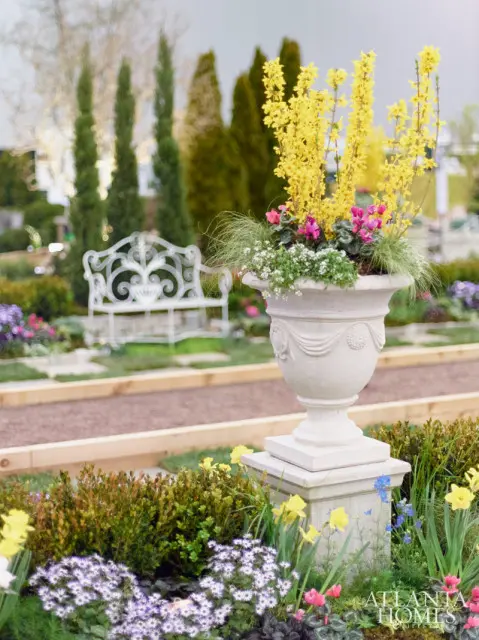 Stone urn overflowing with yellow forsythia and pink cyclamen sits in a garden with a white ornate bench and manicured trees in the background.