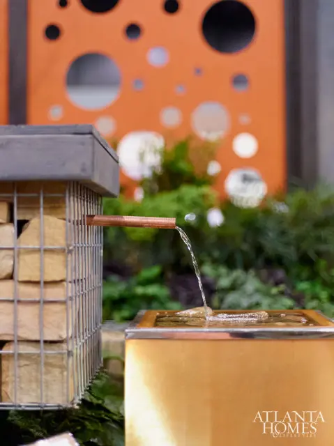 Modern water feature with a gabion wall filled with stones, a copper pipe spout, and a brass basin, set against an orange perforated screen and greenery.