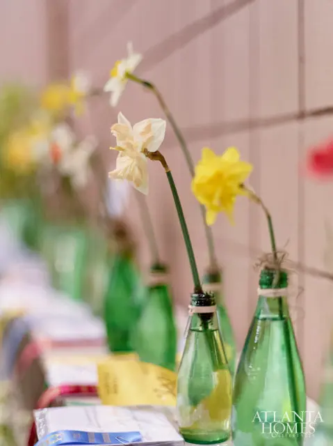 Close-up of daffodils in green glass bottles with a pink wall in the background. The Atlanta Homes & Lifestyles logo is visible in the lower right corner.