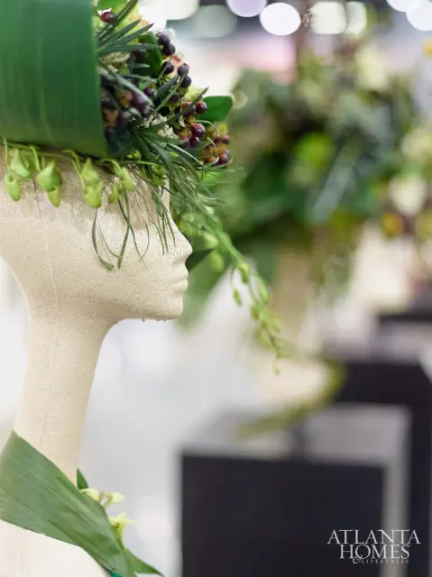 Mannequin head displaying a floral arrangement with dark berries and green foliage, Atlanta Homes & Lifestyles logo in the lower right corner.