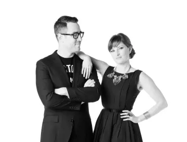 Man in a suit and t-shirt with arms crossed, and woman in a black dress, statement necklace, and striped tights, posing against a white background.