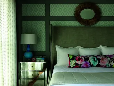 Bedroom with a king-size bed featuring a tall upholstered headboard, white bedding, and a floral bolster pillow. A mirrored nightstand holds a blue lamp and decorative items. The wall has gray paneling with light gray patterned wallpaper inserts and a sunburst mirror above the headboard.