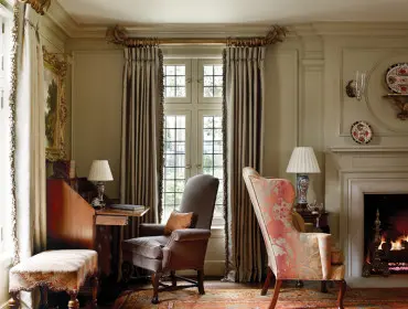 Traditional living room with a fireplace, a wooden writing desk, two armchairs, and floor-to-ceiling drapes in muted tones.
