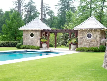 Stone pool house with circular windows and a wooden pergola connects to a second structure. A bright blue swimming pool sits in front of a manicured lawn and lush trees.