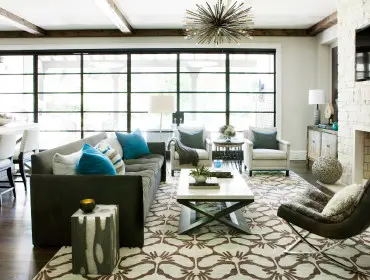 Living room with dark gray tufted sofa, two cream armchairs, a white coffee table, and a stone fireplace. Large windows overlook an outdoor patio.