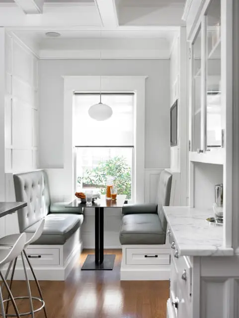 Kitchen banquette with gray tufted upholstery and built-in white drawers, next to a window with a view of greenery.