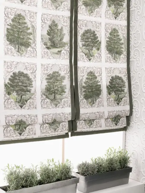 Roman shades with a toile pattern of trees and ornate scrollwork, trimmed in dark green fabric, hang above a window with planters of rosemary.