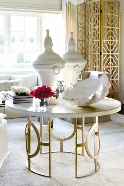 Living room with a round marble coffee table featuring a gold geometric base, topped with a vase of red roses, books, and a large white shell.