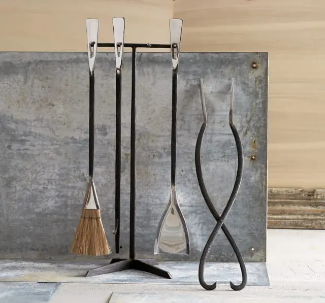 Black metal fireplace tool set with brushed nickel accents, including a broom, shovel, poker, and tongs, displayed against a rustic galvanized metal backdrop.