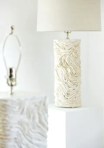 Close-up of a textured white ceramic table lamp with a linen shade, set on a white pedestal. Another lamp base is visible in the blurred background.