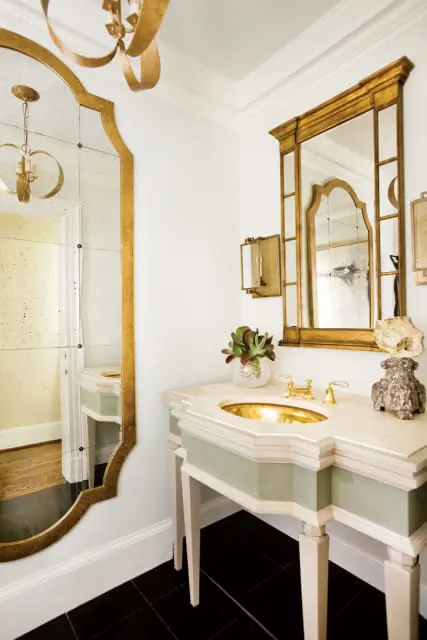 Powder room with a gold-framed antique mirror, a cream vanity with a gold sink and brass fixtures, and black tile flooring.