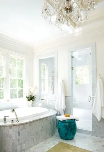 White bathroom with a large soaking tub, marble tile accents, glass-enclosed shower, and a crystal chandelier.