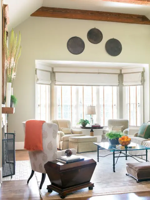 Living room with pale green walls, exposed wooden beams, and a bay window with white roman shades. Furnishings include upholstered chairs, a glass coffee table, and a dark wood chest.