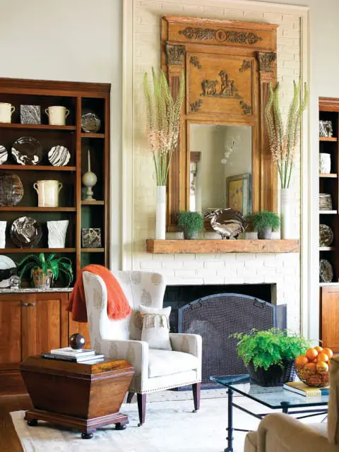 Living room with a white brick fireplace, ornate wooden mirror, built-in wooden bookshelves, and a patterned armchair.