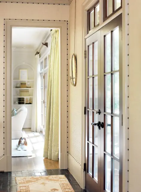 Hallway with textured wallpaper, a double-pane glass door, and a view into a sunlit room with a dog on a rug and built-in shelves.