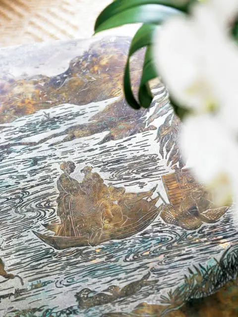 Close-up of an antique metal tray with an engraved scene of boats on water, featuring figures in traditional clothing and ducks swimming.