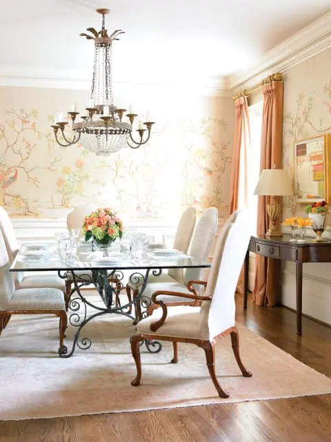 Dining room with a glass-top table, ornate metal base, upholstered chairs, a crystal chandelier, and wallpaper with a bird and floral motif.
