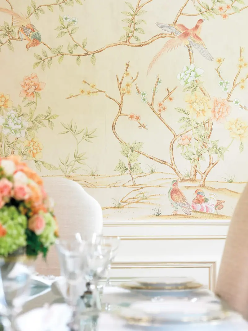 Dining room with Chinoiserie wallpaper featuring birds and flowers, upholstered chairs, and a table set with china and crystal glasses.