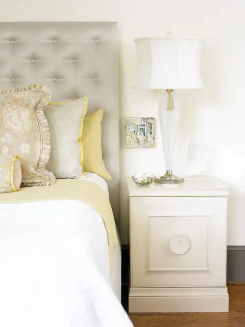 Bedroom with a tufted gray headboard, white bedding, yellow accent pillows, and a cream nightstand with a white lamp.