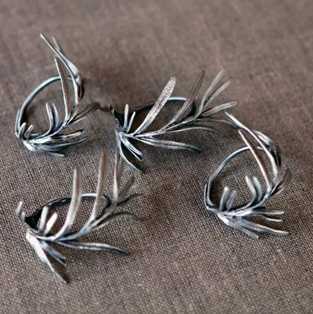 Four silver napkin rings shaped like rosemary sprigs on a textured brown fabric background