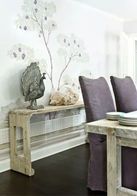 Dining room with a wall mural of trees with purple fruit, a distressed wood console table with a peacock sculpture, and upholstered chairs with a rustic wood table.