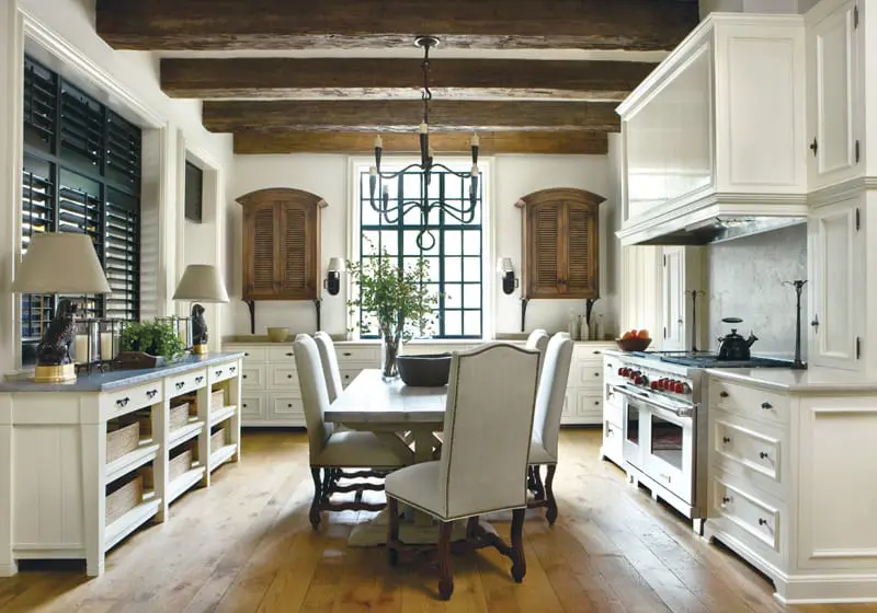 Cream kitchen with white custom cabinetry, exposed wooden ceiling beams, a large window with black grid panes, and a dining table with upholstered chairs.