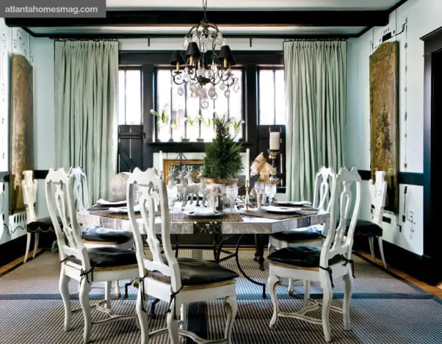Dining room with a large octagonal table set for a meal, surrounded by ornate white chairs with black cushions. Light blue walls, dark wood beams, and a crystal chandelier complete the space.