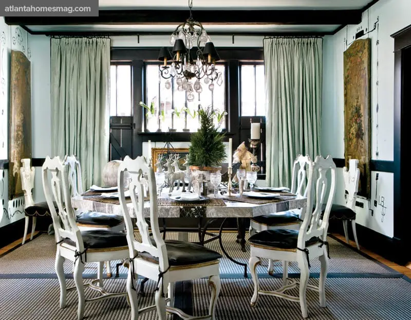 Dining room with a large octagonal table set for a meal, surrounded by ornate white chairs with black cushions. Light blue walls, dark wood beams, and a crystal chandelier complete the space.