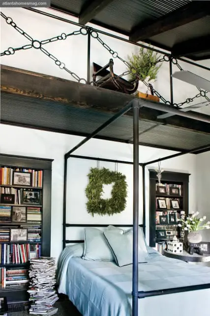 Bedroom with a black metal canopy bed, light blue bedding, and two bookshelves filled with books and photos. A square green wreath hangs on the wall above the bed.