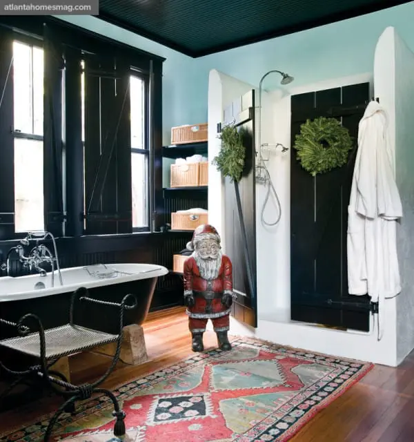 Bathroom with clawfoot tub, black shutters, open shower with barn doors, woven baskets on shelves, and a Santa figurine on a colorful rug.
