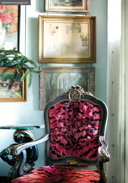 Ornate armchair upholstered in pink and black tiger-print fabric, with several framed paintings on a light blue wall behind it.