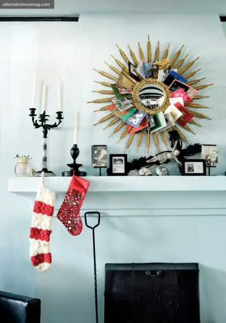 Light blue living room with a white mantelpiece decorated for Christmas, featuring two stockings, framed photos, and a sunburst mirror adorned with holiday cards.