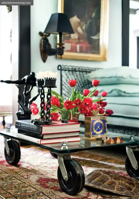 Glass coffee table with industrial wheels, stacked books, red tulips, a brass clock, and a black sculpture.