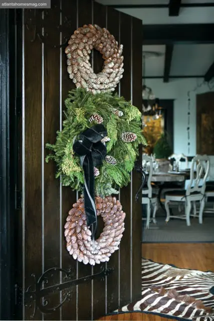 Dark wood door with three wreaths: two made of pinecones and one of evergreen branches with a black velvet bow. A zebra print rug is visible on the floor.