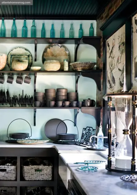 Kitchen with light blue walls, dark wood shelves displaying blue glass bottles, ceramic dishes, dark gray mugs, and stacks of plates. A marble countertop features baskets and a vintage hourglass.