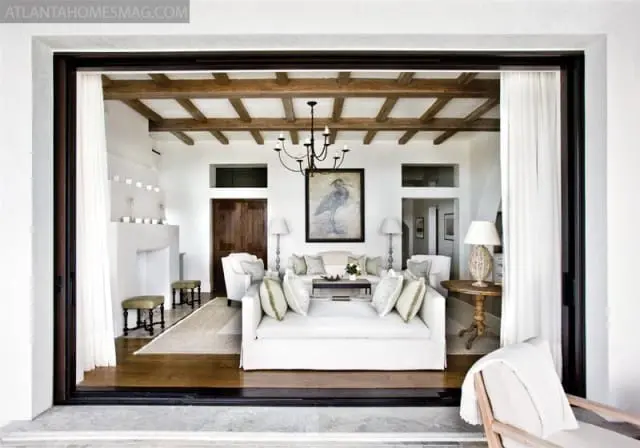 Living room with white slipcovered sofas, exposed wooden ceiling beams, a large painting of a bird, and a dark wood door.