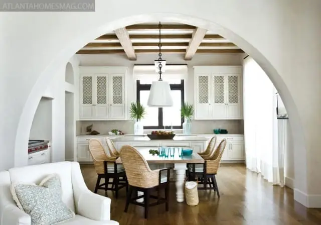 White kitchen with beamed ceiling, white cabinetry with lattice doors, and a dining table with woven chairs.