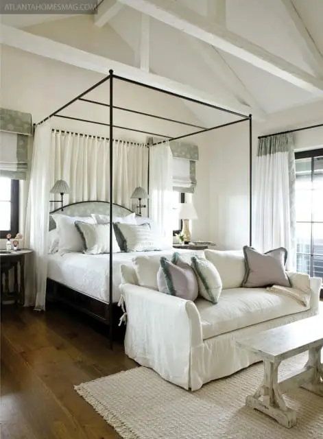 Bedroom with a dark metal four-poster bed draped in white sheer curtains, a white slipcovered sofa with decorative pillows, and exposed white beams on the vaulted ceiling.