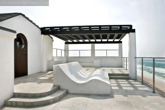White stucco building with dark wood door and circular window, leading to a patio with a modern concrete chaise lounge and a black pergola overlooking the ocean.