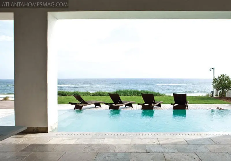 Infinity pool overlooking the ocean with four dark lounge chairs on a wooden deck, framed by a white architectural element.