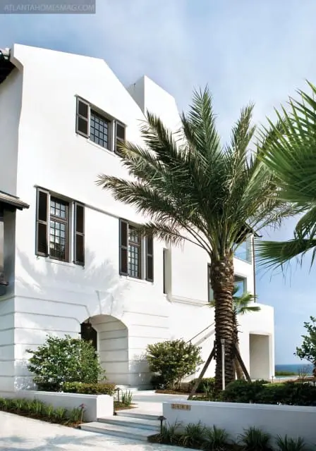 White stucco house with dark shutters on windows, arched entryway, and palm trees in front.