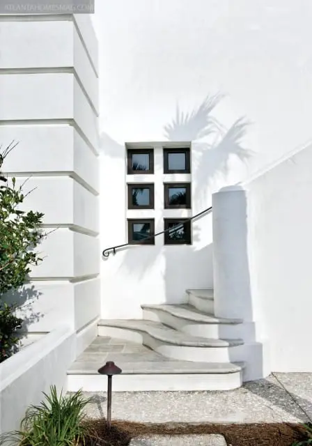 White stucco exterior with a curved stone staircase, a dark-framed window with six panes, and palm frond shadows on the wall.