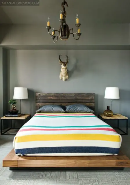 Bedroom with gray walls, a wooden platform bed with a striped blanket, two nightstands with lamps, and a taxidermied antelope head mounted on the wall.