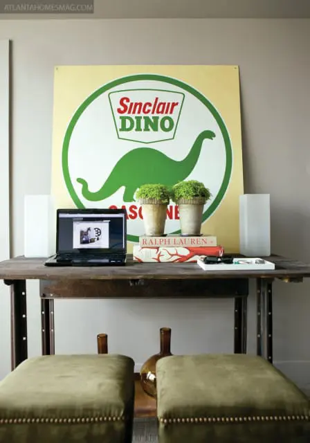 Desk with laptop, books, and two potted plants in front of a large vintage Sinclair Dino gasoline sign.