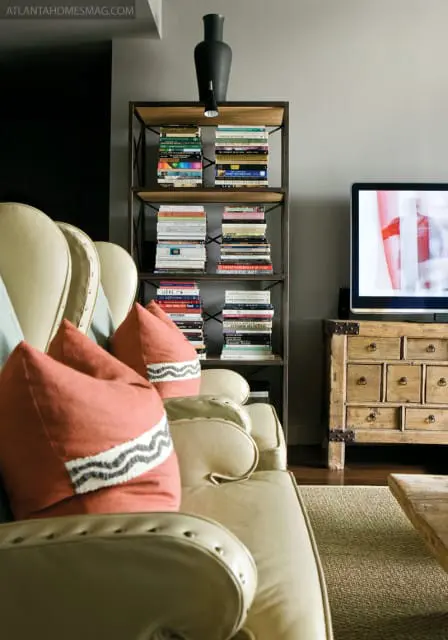 Living room with a cream leather sofa, coral throw pillows with black and white zigzag trim, a metal bookshelf filled with books, and a wooden chest of drawers with a television on top.